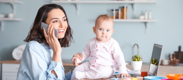 A smiling mom talks on a mobile phone. A baby sits on a desk beside her. 